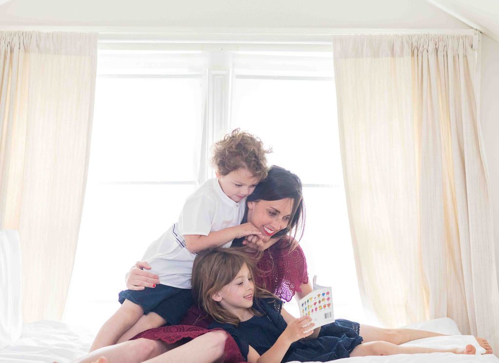 Woman laying on a bed with her two kids. She is wearing cranberry eyelet top and skirt. The young boy is wearing a white popover with blue embroidery. The little girl is reading and wearing a navy swing dress with lace inserts and flutter sleeves. 