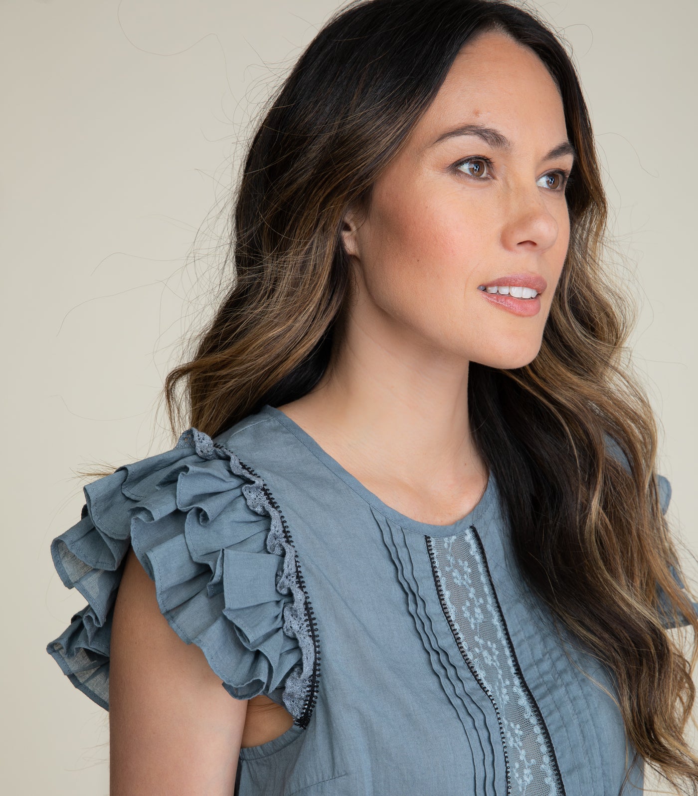 Image of a brunette looking off camera and wearing a blue gray cotton top with lace inserts, pintucks, and flutter sleeves. 