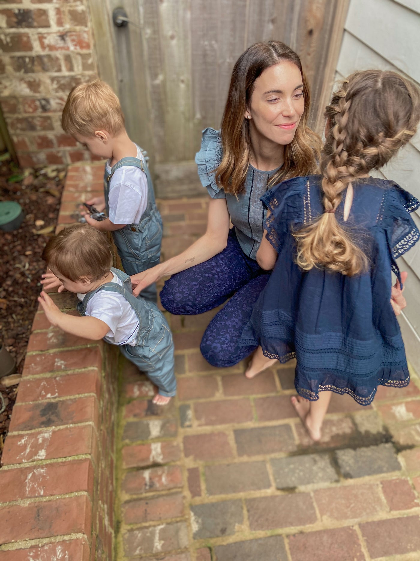 Woman wearing a blue shirt with lace insert and navy embroidered joggers is kneeling down with her three kids outsides. The boys are in blue embroidered overalls. The girl has her hair braided and is in a navy dress with lace insert and pintucks.  