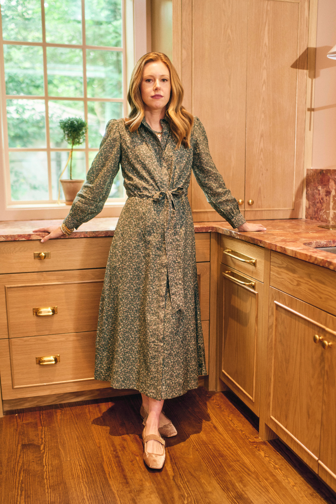 Woman in a patterned dress standing in a kitchen with wooden cabinets and a window.