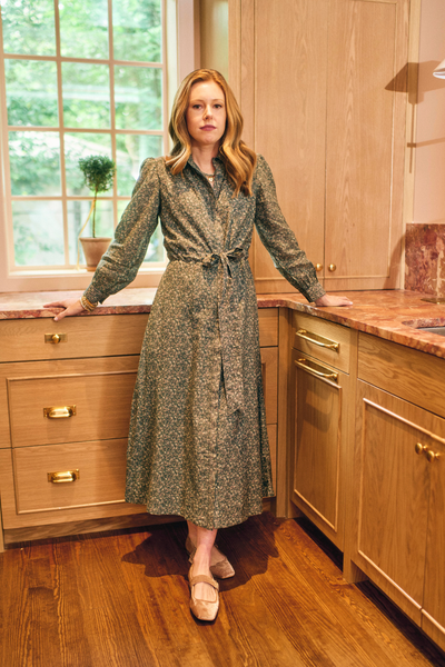 Woman in a patterned dress standing in a kitchen with wooden cabinets and a window.