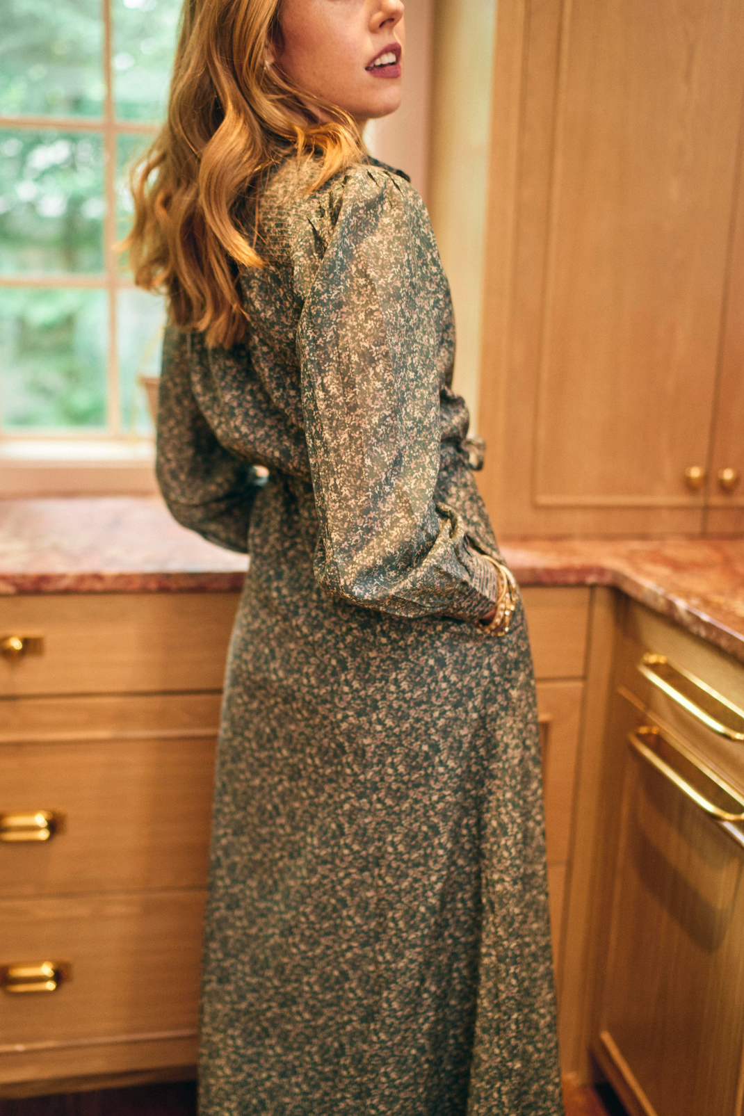 Woman in a patterned dress standing in a kitchen with wooden cabinets and marble countertops.