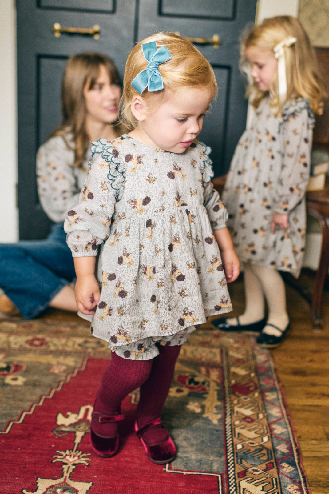 Two young girls in matching dresses standing on a patterned rug with a woman in the background.