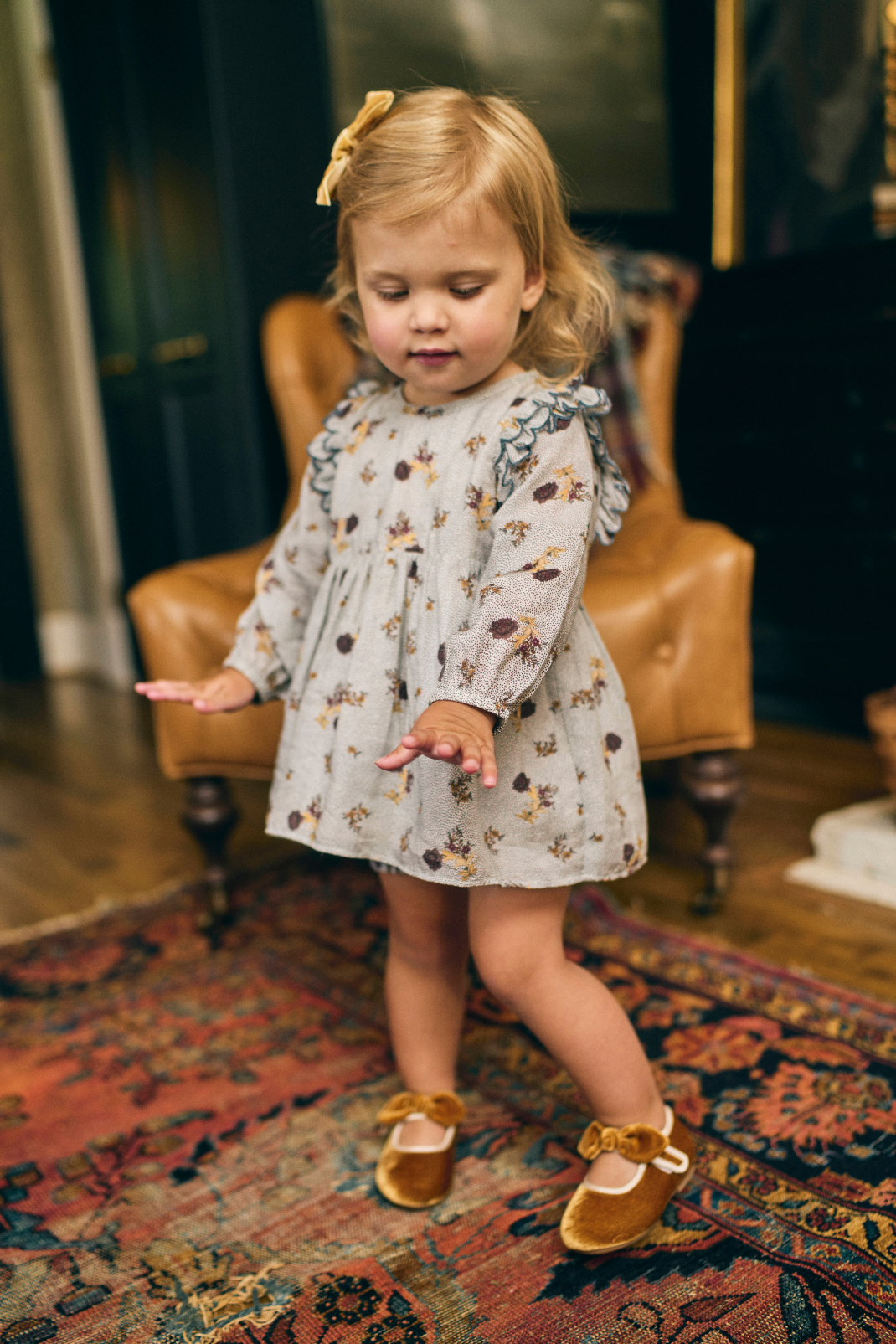 Young girl in a floral dress standing on a patterned rug in a room with a brown leather chair.
