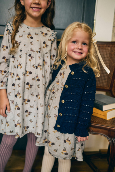 Two young girls standing indoors, one in a floral dress and the other in the same floral dress with a navy cardigan.