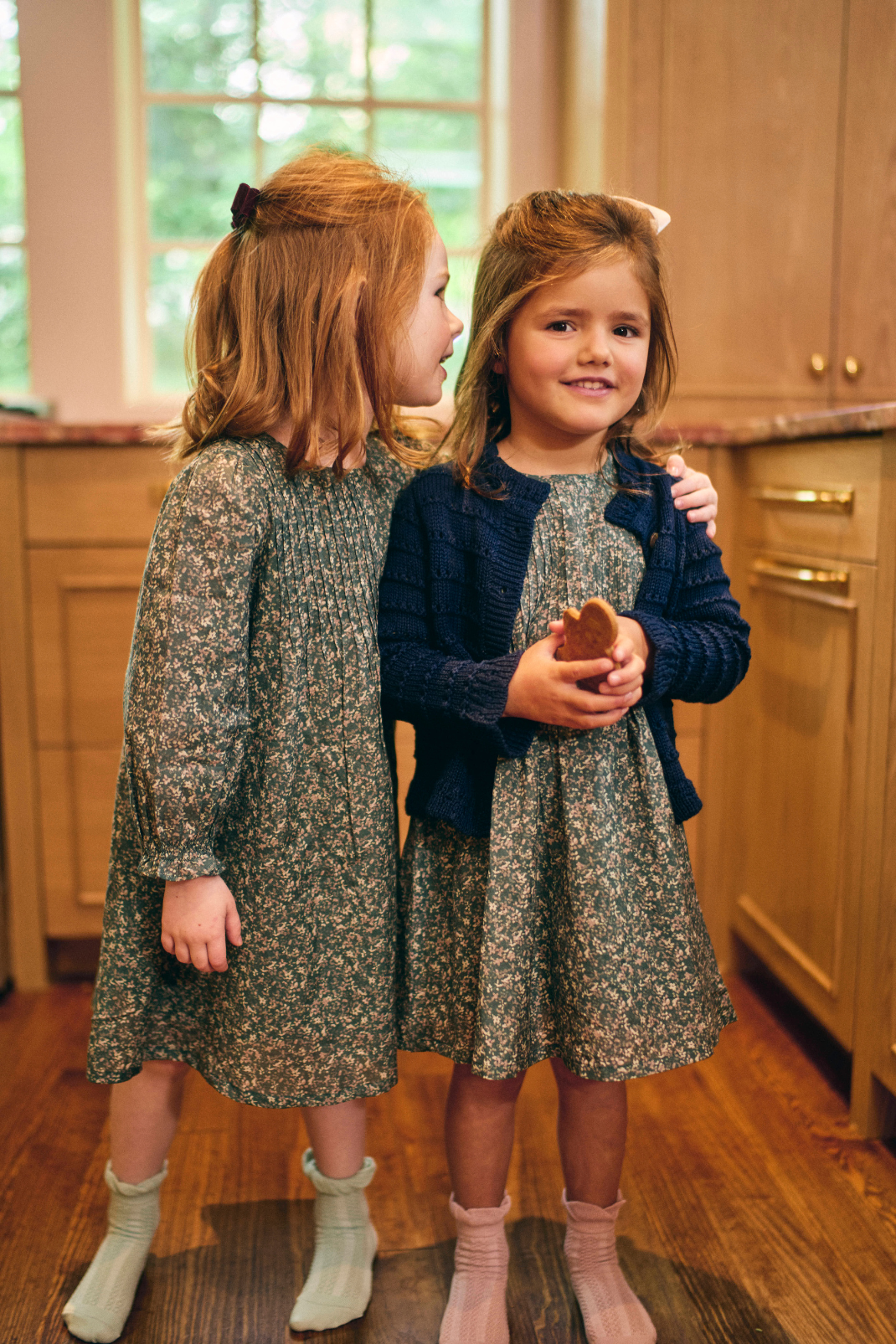 Two young girls in matching dresses standing in a kitchen.