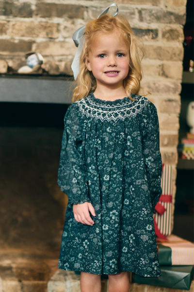 Young girl in a green floral dress standing in front of a stone fireplace.