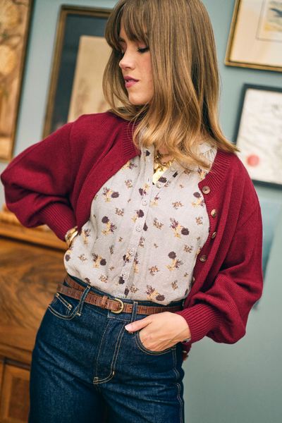Woman wearing a red cardigan over a floral blouse with blue jeans indoors.
