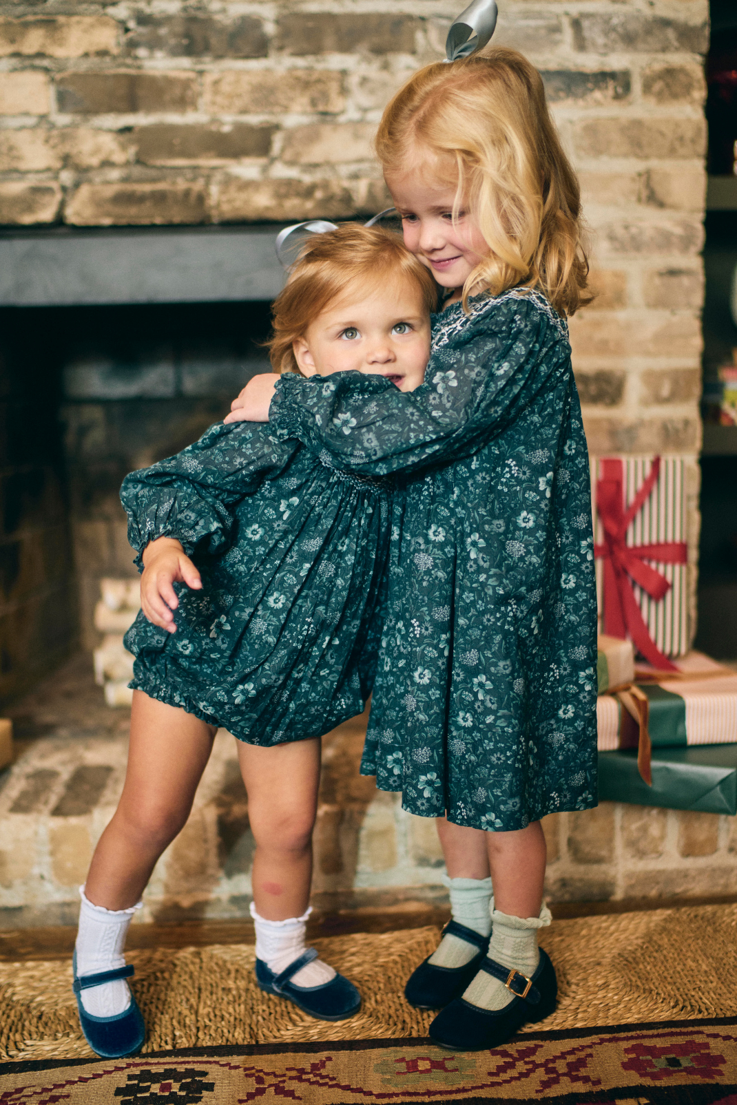 Two young girls in matching green floral dresses standing in front of a stone fireplace.