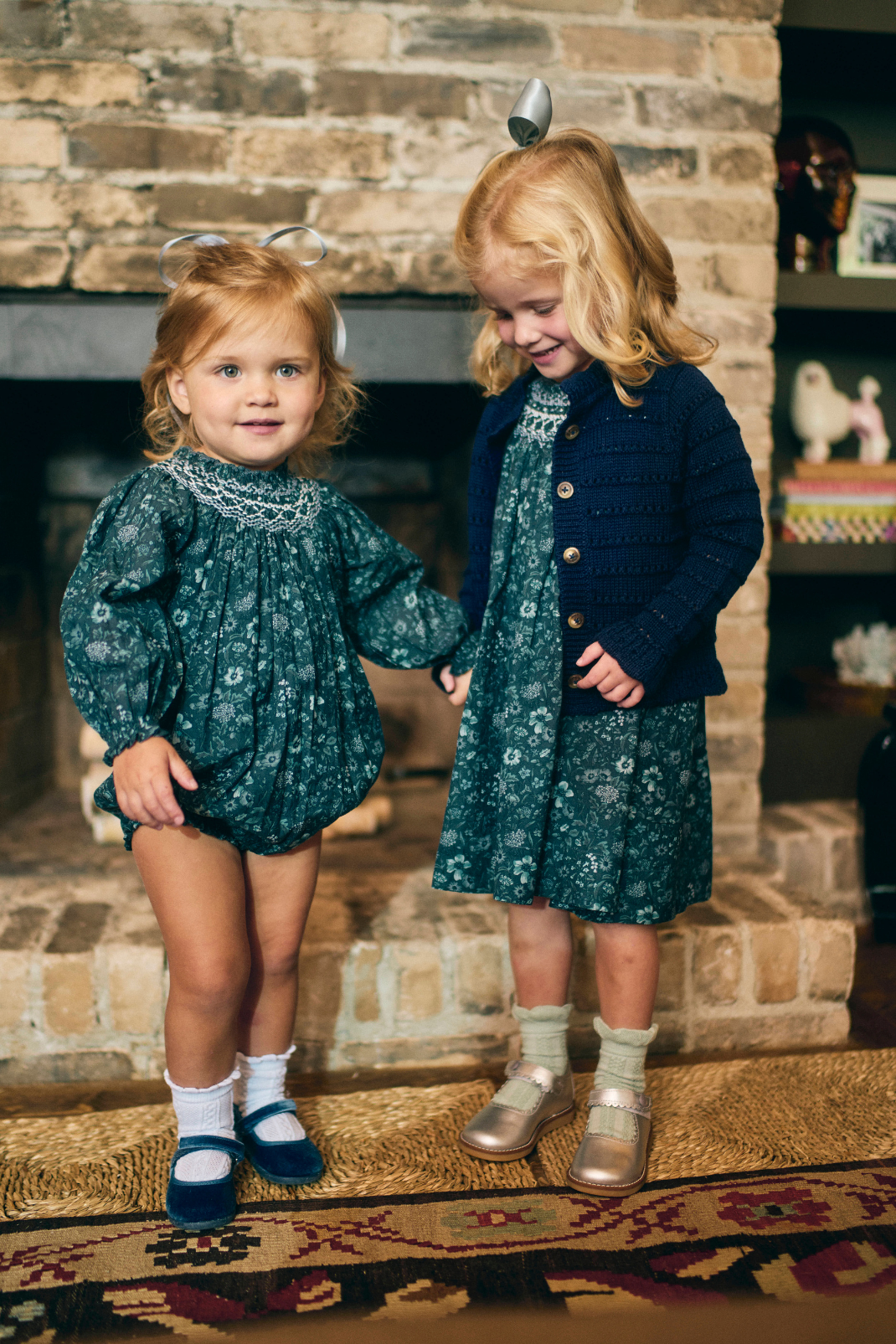 Two young girls in matching outfits standing in front of a fireplace.