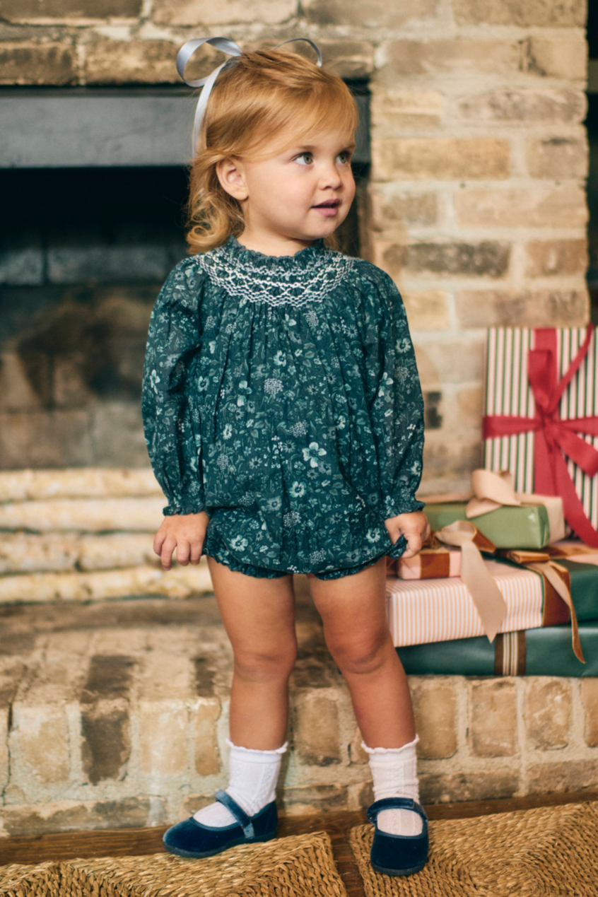 Young child in a floral outfit standing in front of a fireplace with gift boxes.