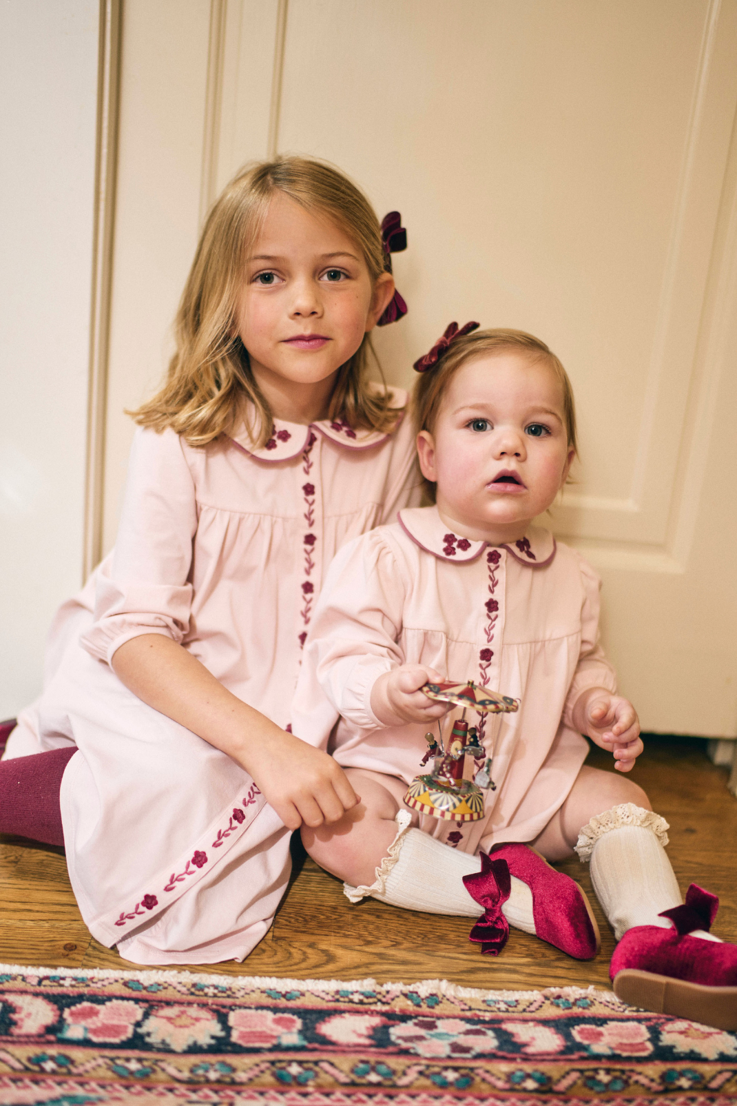 Two young girls in matching pink dresses sitting on a patterned rug.
