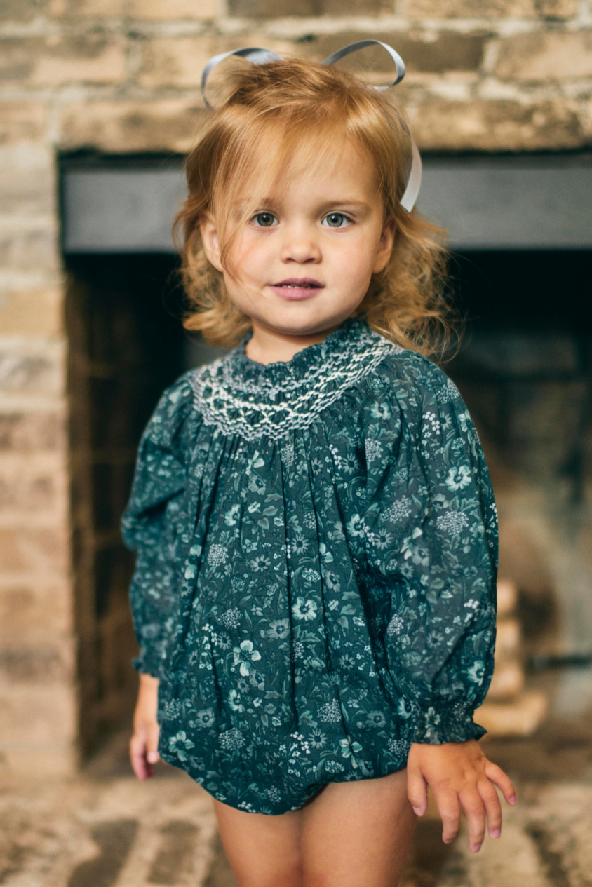 Young girl in a floral dress standing in front of a stone fireplace.