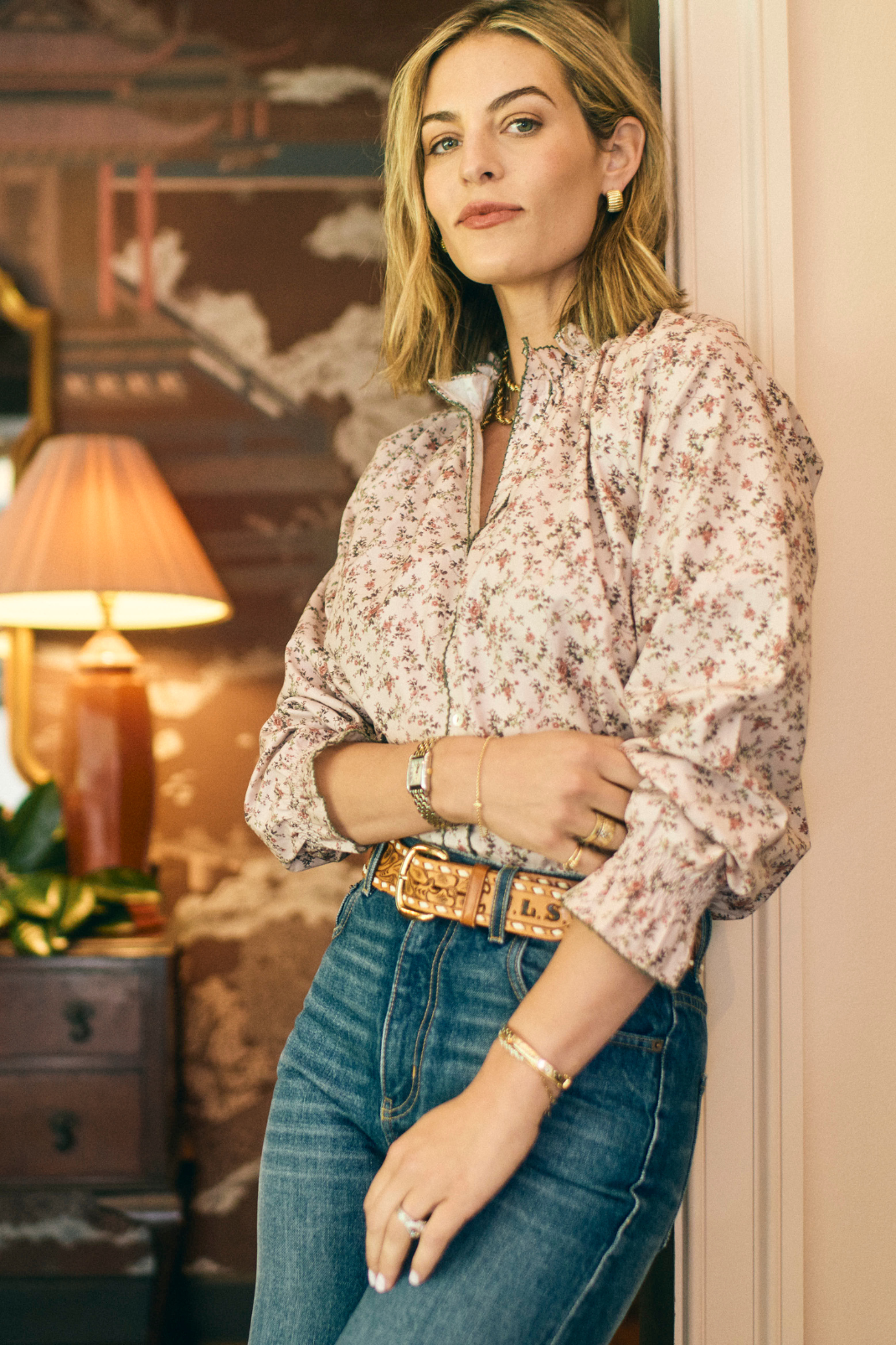 Woman wearing a floral blouse and jeans in a room with a lamp and decorative wall.