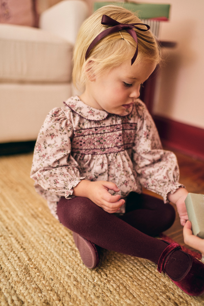 Young girl in a floral dress and maroon tights sitting on the floor.