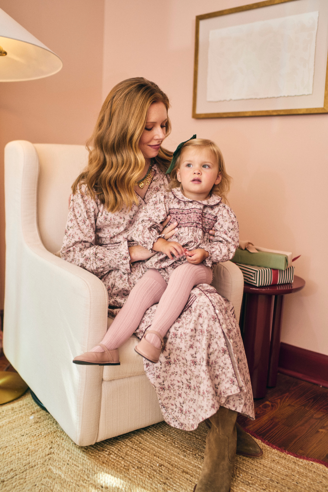 Woman and child in matching floral dresses sitting on a white chair in a room with pink walls.