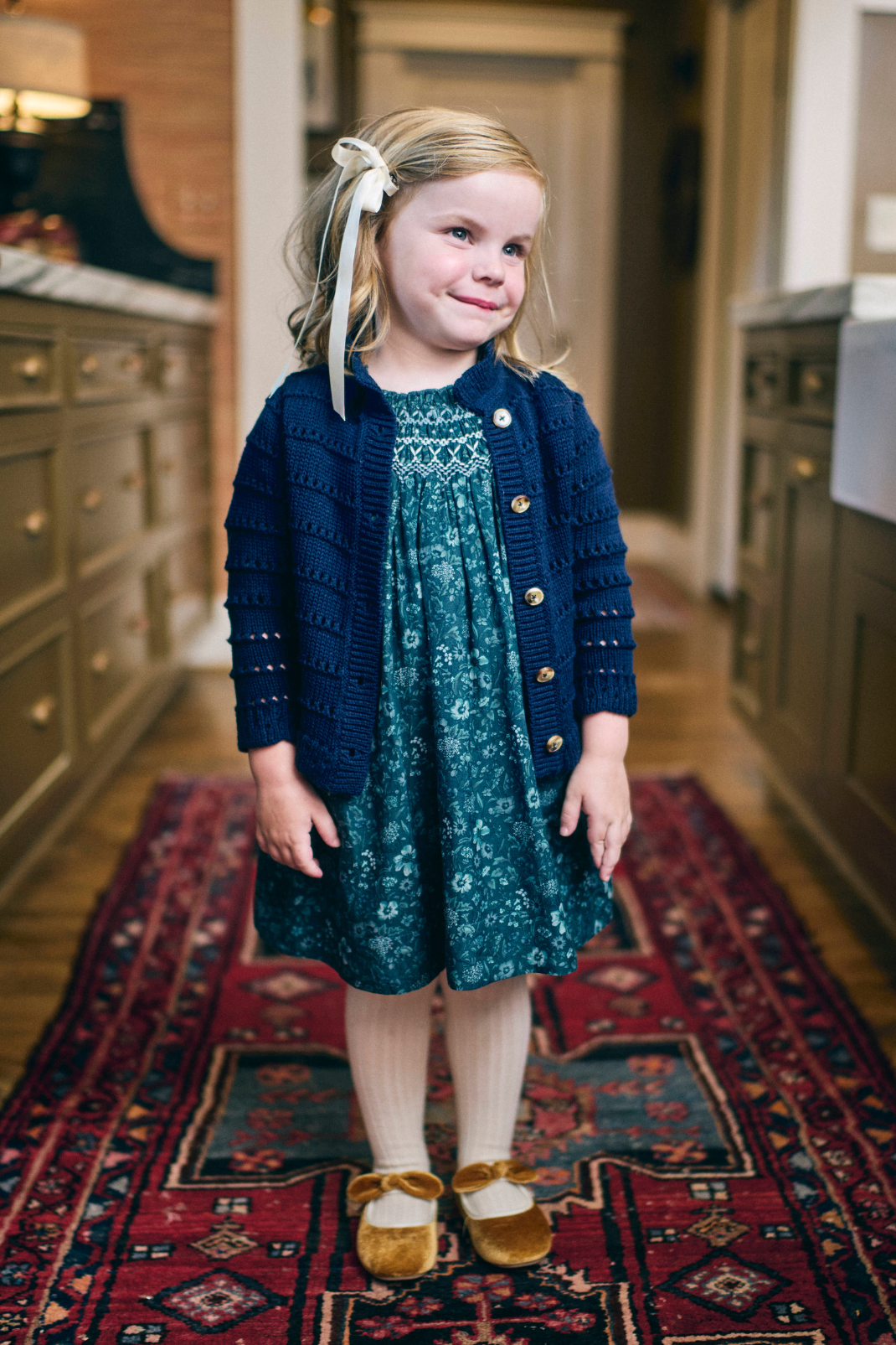 Young girl in a blue cardigan and teal dress standing on a patterned rug.