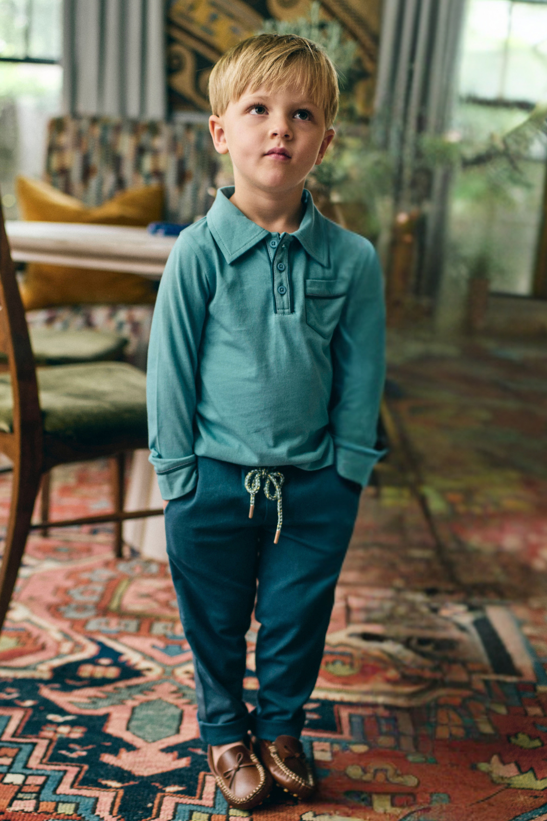 Young boy in teal shirt and navy pants standing on a patterned rug in a room with furniture and plants.