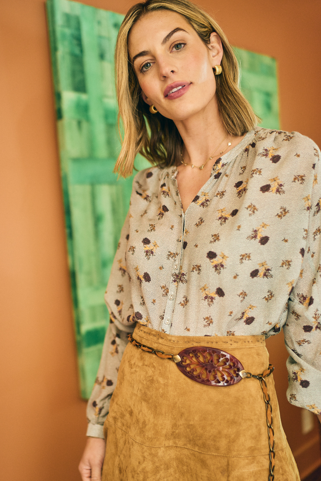 Woman wearing a floral blouse and brown skirt with a decorative belt, standing against an orange wall with a green abstract painting.
