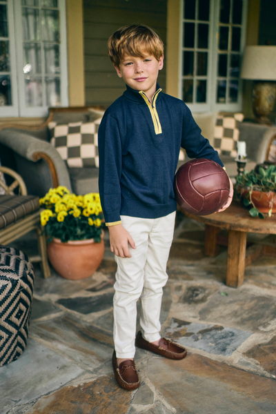 Young boy holding a soccer ball on a stone patio wearing a navy sweater with a golden yellow zippered placket
