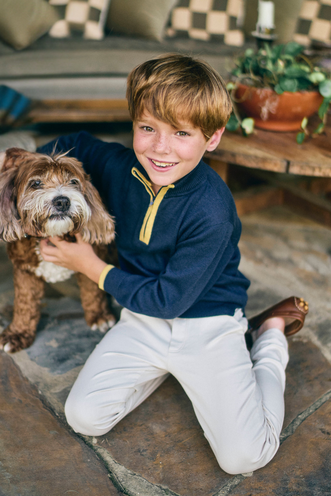 Young boy sitting on a stone floor with a dog, wearing a blue sweater and white pants, in a cozy indoor setting.