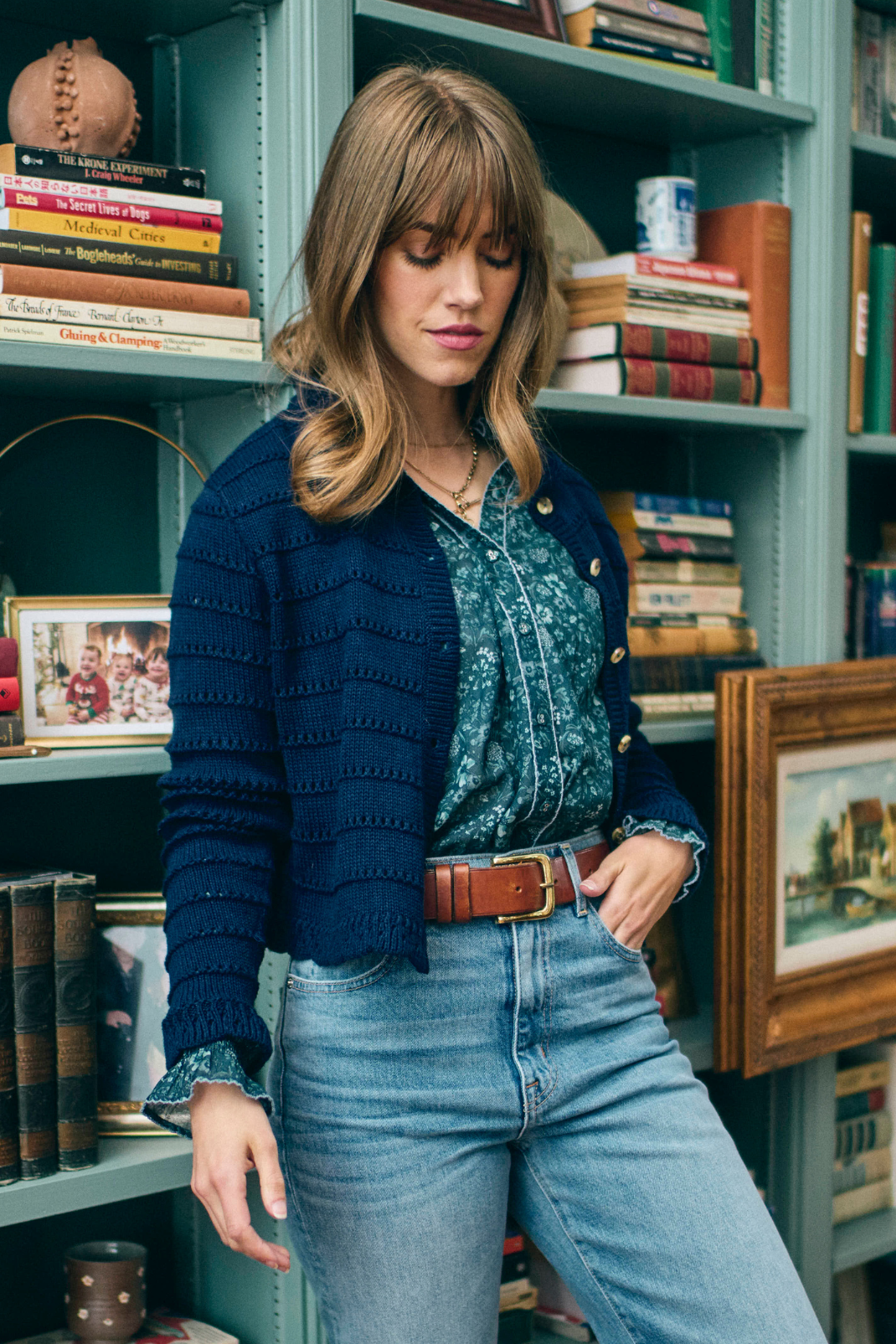 Woman standing in a blue cardigan and blue floral top next to bookshelves and books in the background