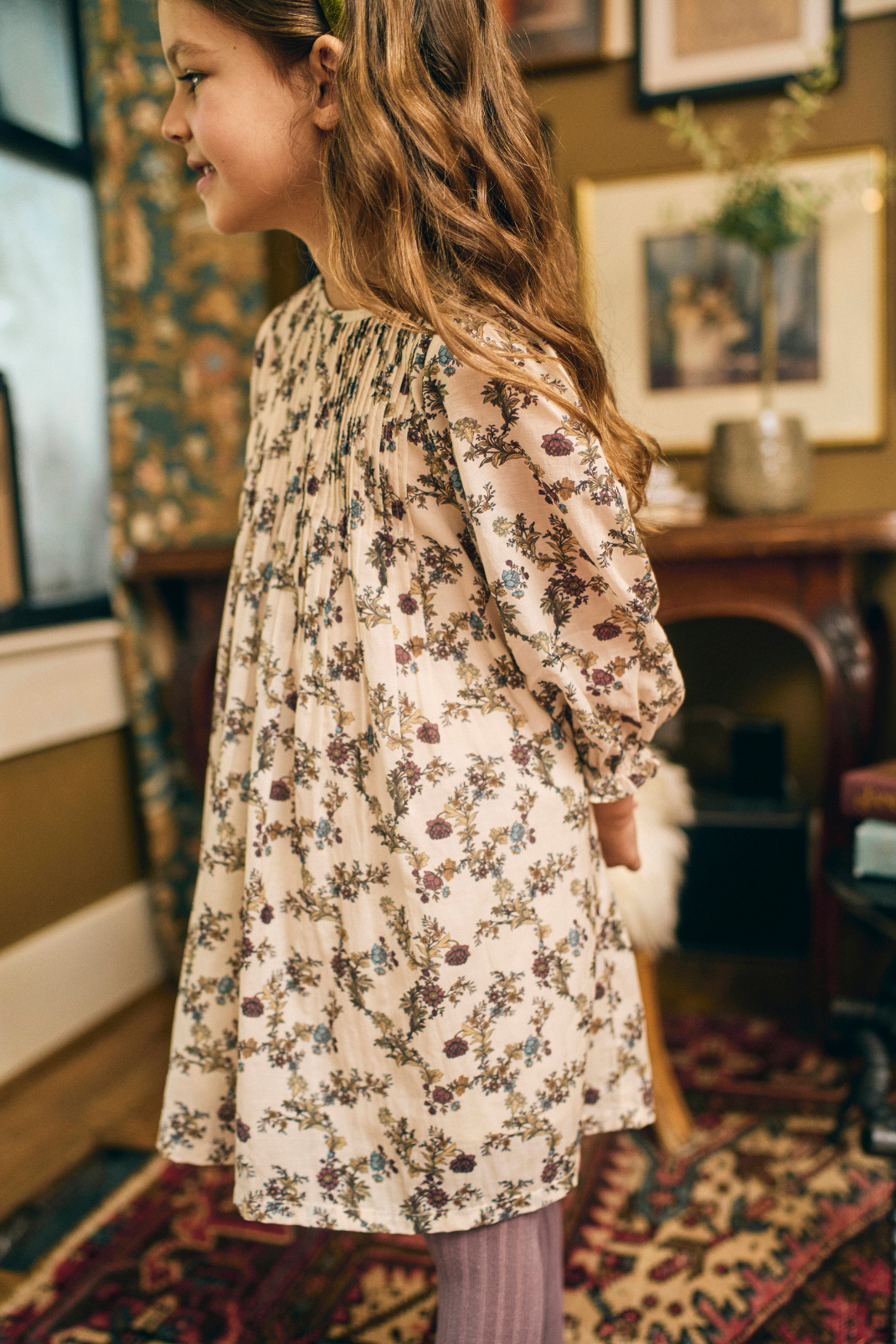 Young girl wearing a floral dress in a room with a patterned rug and framed pictures on the wall.