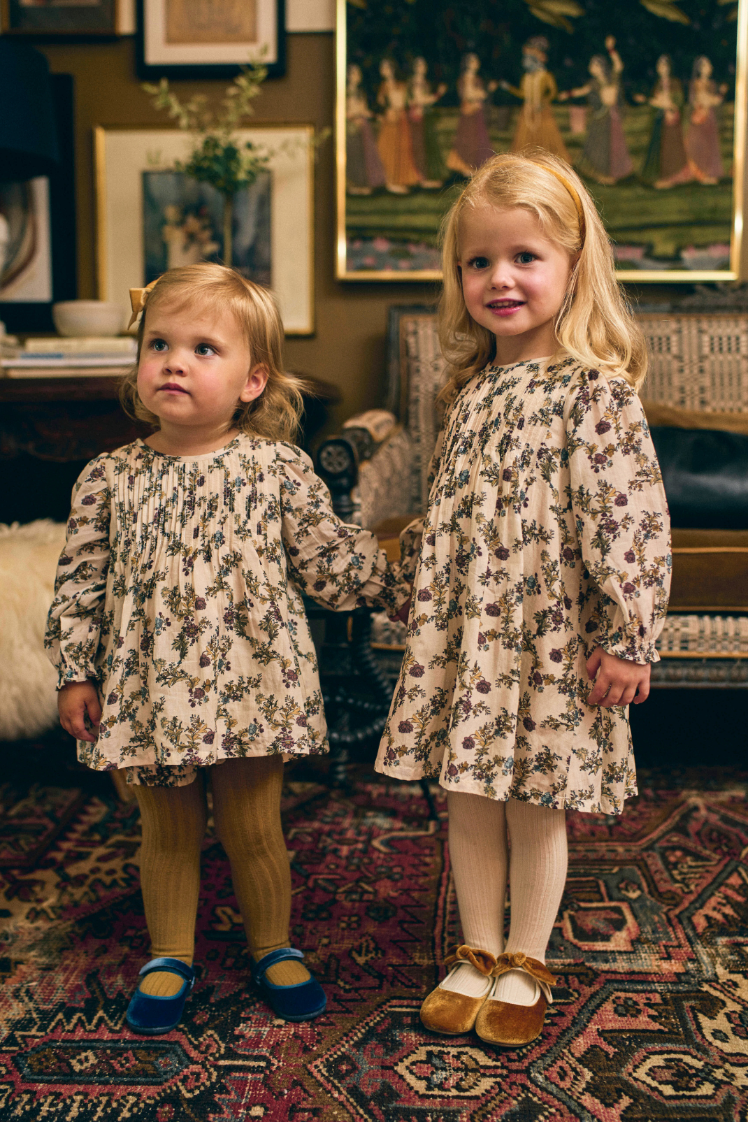 Two young girls in floral dresses standing in a room with vintage decor.