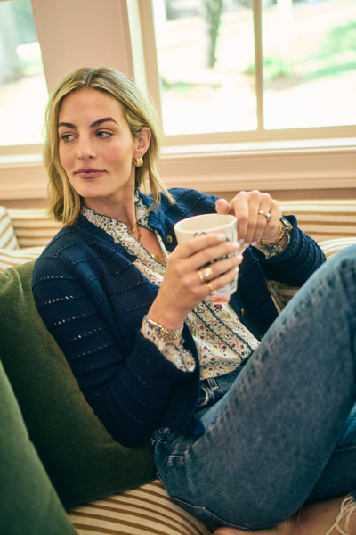 Woman sitting on a couch in a blue sweater and a floral printed shirt holding a mug in a cozy indoor setting