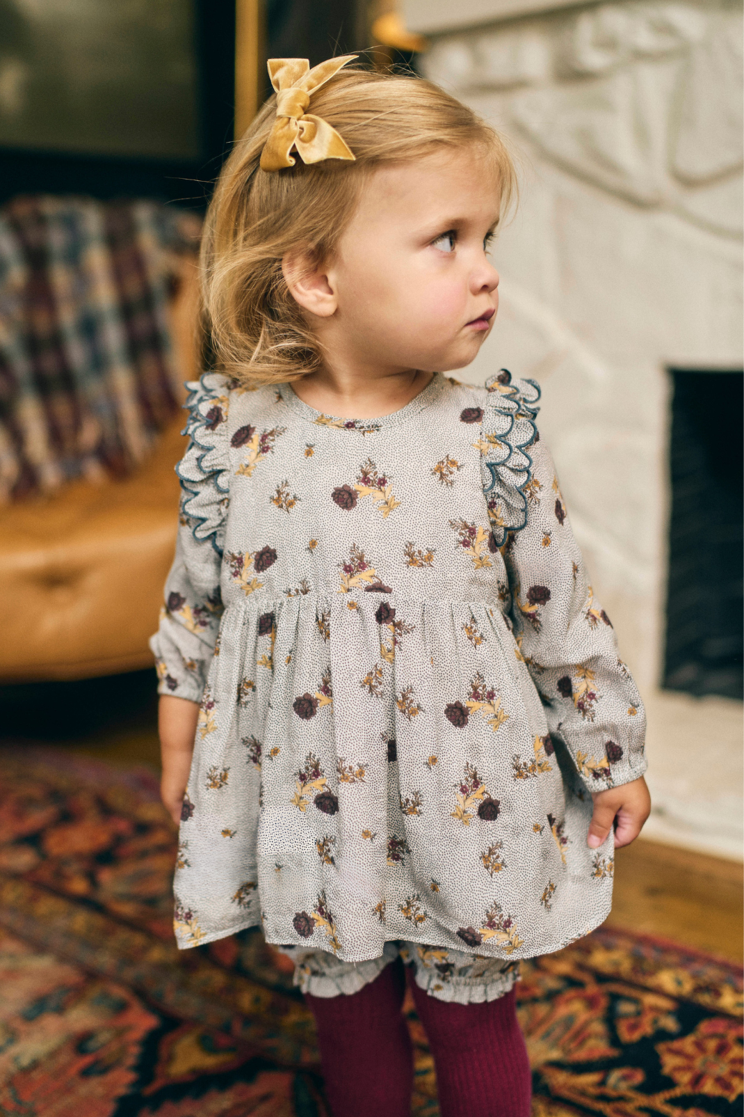 Young girl in a floral dress standing in a room with a fireplace.