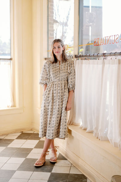 A woman standing next to a window wearing the Mallie Dress in Grey Gardens