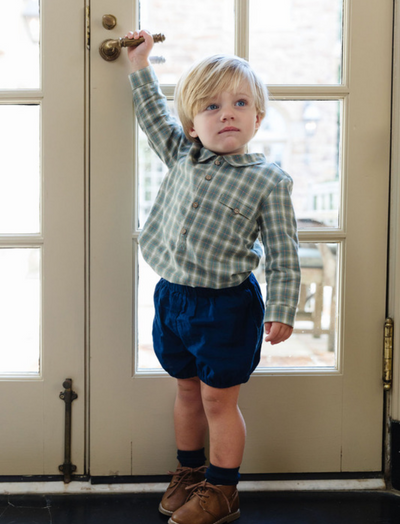 Little boy wearing a long sleeve checkered shirt and blue shorts trying to open a door and go outside.