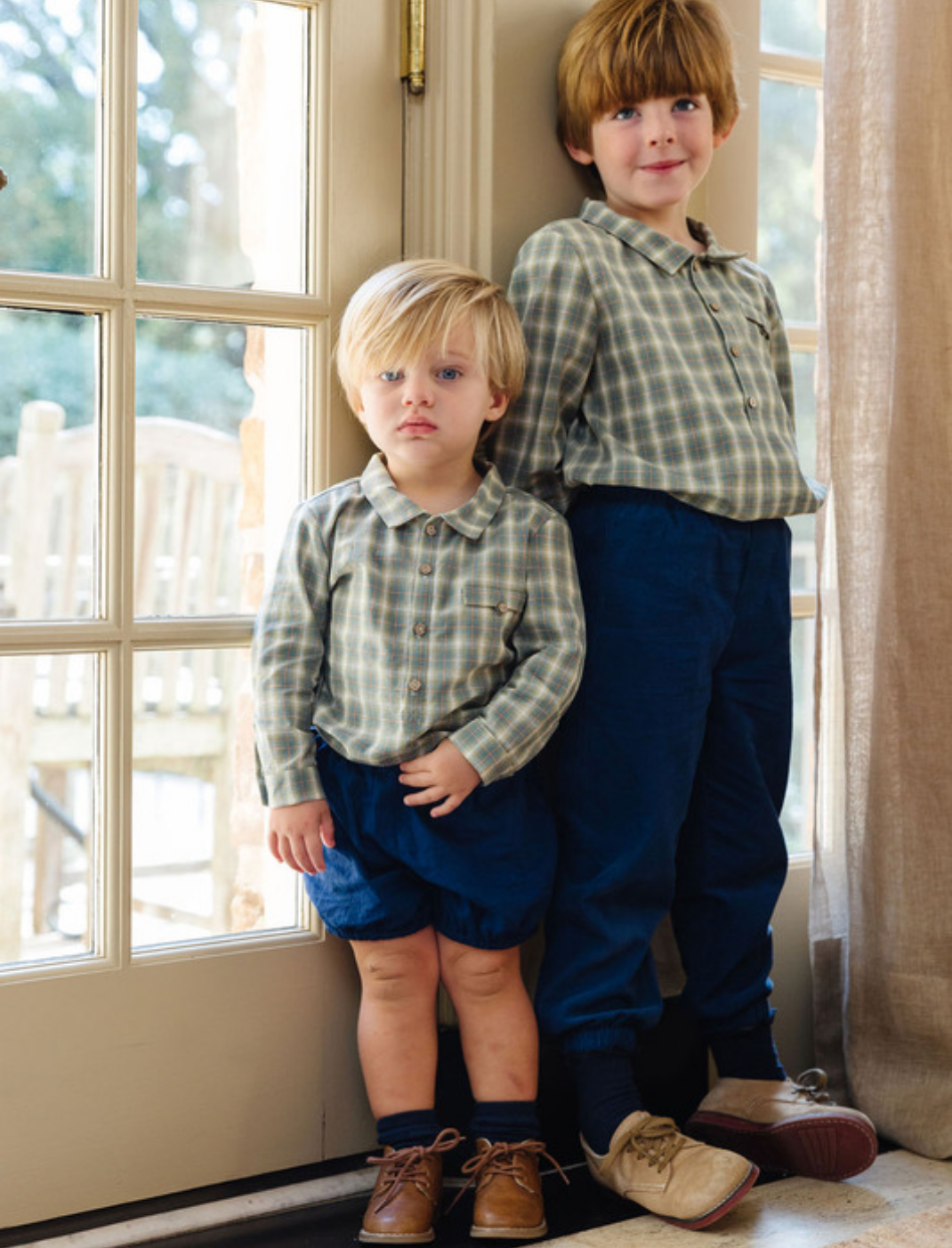 Two young boys leaning against a wall next to a windowed door inside. Both boys wearing a long sleeve checkered shirt and the older boy is wearing blue pants and the younger boy is wearing blue shorts.